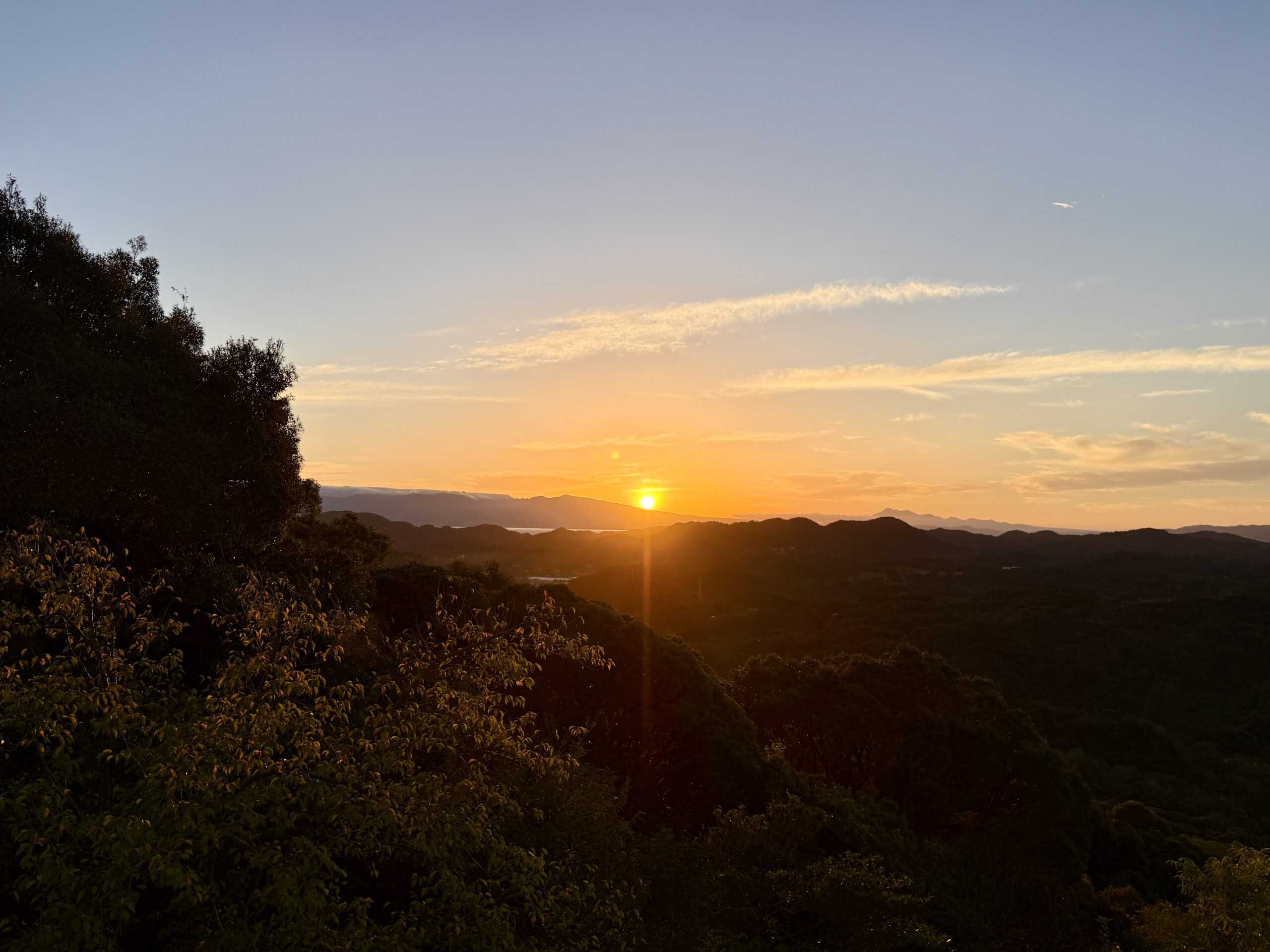 東の稜線から昇る朝日と、木立のシルエットが重なる風景。秋晴れの空に一条の光が差し込む。
